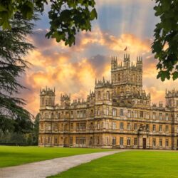 The yellow stone exterior of Highclere Castle with path and lawn in foreground and sun lit clouds above.