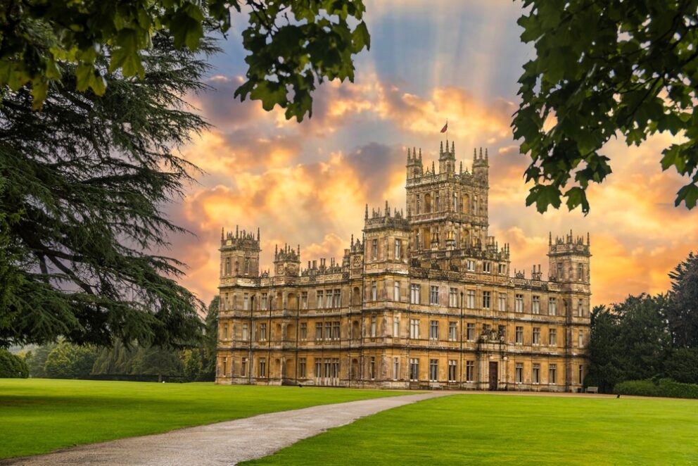 The yellow stone exterior of Highclere Castle with path and lawn in foreground and sun lit clouds above.