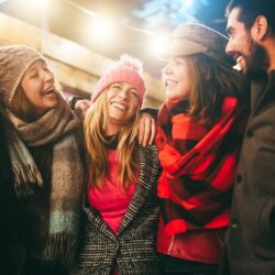 A group of five adult friends, dressed in winter coats, scarves and hats, laughing together.