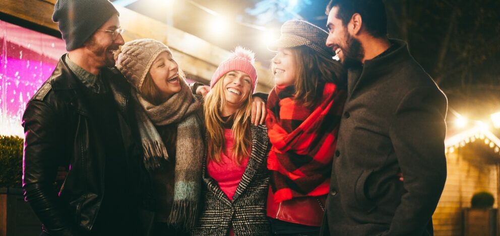A group of five adult friends, dressed in winter coats, scarves and hats, laughing together.