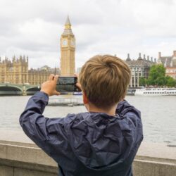 Child in blue raincoat looking across the River Thames, photographing Big Ben on their phone.