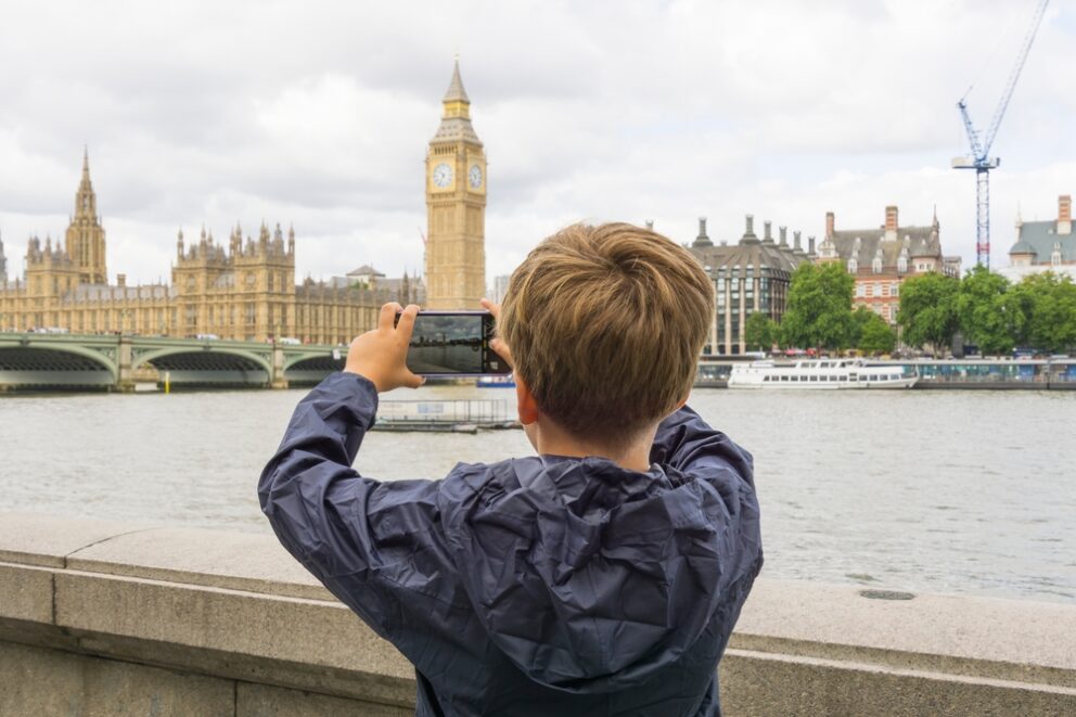 Child in blue raincoat looking across the River Thames, photographing Big Ben on their phone.
