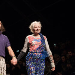 Vivienne Westwood on the catwalk holding hands with another figure, with a black background.