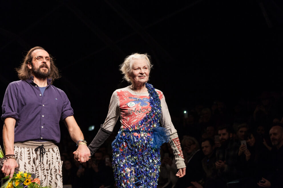 Vivienne Westwood on the catwalk holding hands with another figure, with a black background.