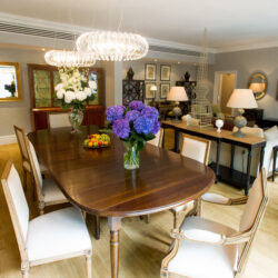 A large living room with dining table and chairs in foreground (dressed with a vase of large blue flowers) and seating area in background.